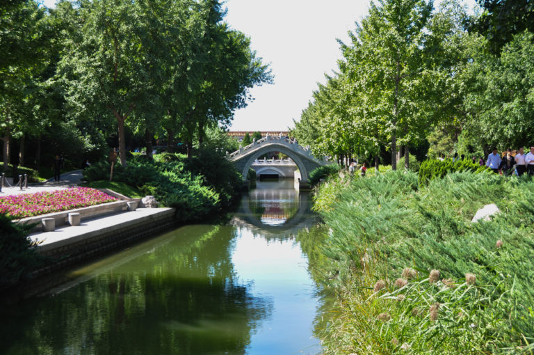 Calm green canal in a Beijing park leading to a distant white moon bridge bordered by dense plantings.