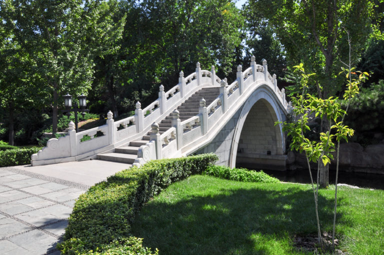 White stone moon bridge with a high single arch over a narrow canal in a landscaped Beijing park.