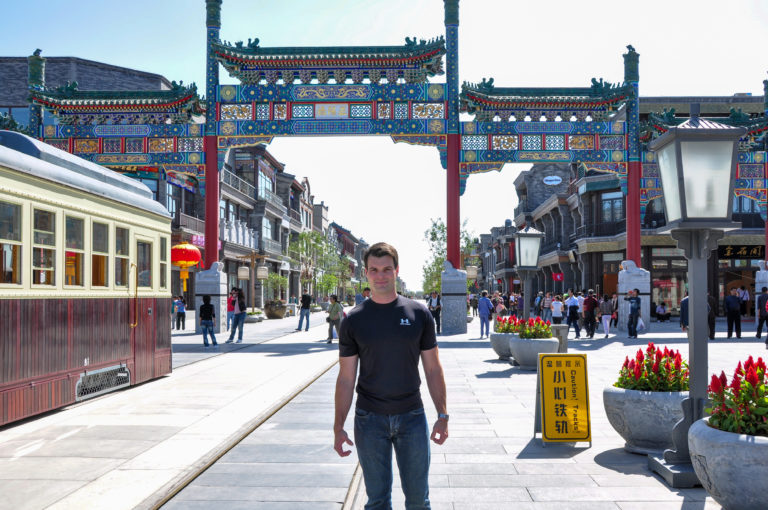 Traveler standing under a colorful paifang beside a red-and-cream heritage tram on Qianmen Street in Beijing.