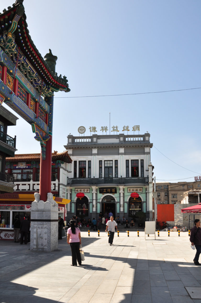 Historic Beijing silk or textile shop with gold Chinese rooftop characters, red lanterns, and arched white facade with green trim in a commercial district.