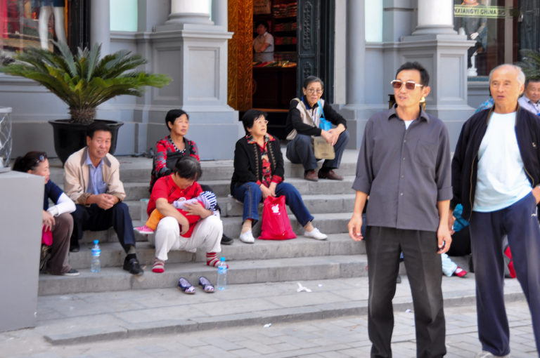 Several adults sit on wide stone steps while two men stand in the foreground outside a public building in Beijing.
