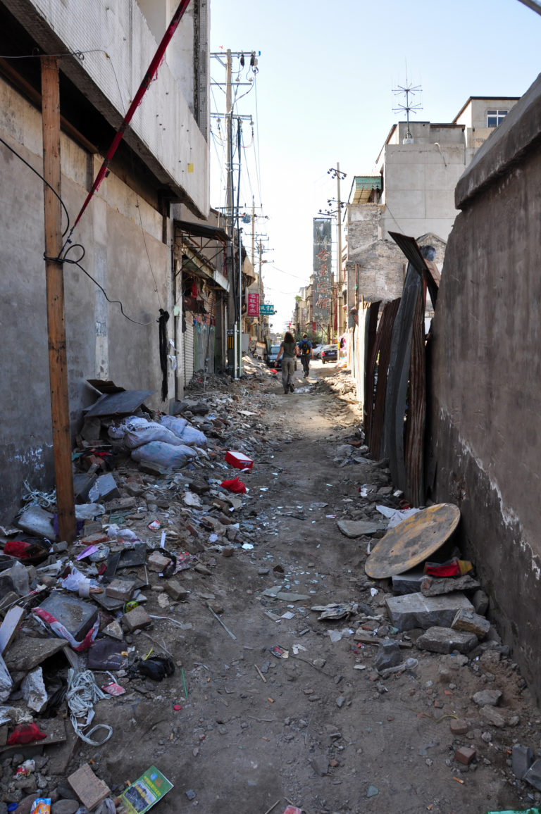 Narrow Beijing hutong alley with partially demolished walls and piles of rubble on both sides.