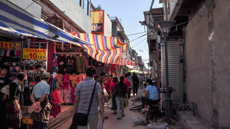 Pedestrians walk through a narrow Beijing hutong market under colorful striped awnings with clothing stalls on both sides.