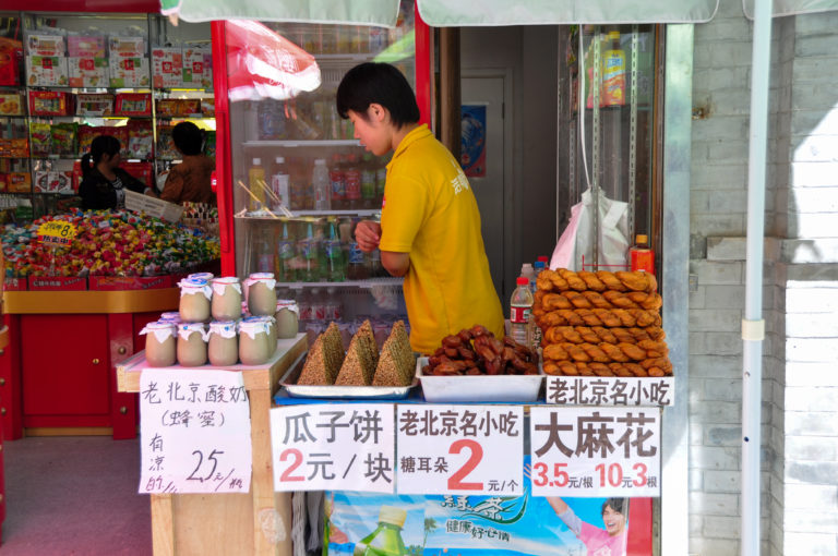 Beijing street-food stall with bottled yogurt, trays of triangular 瓜子饼, and handwritten price signs in Chinese yuan.