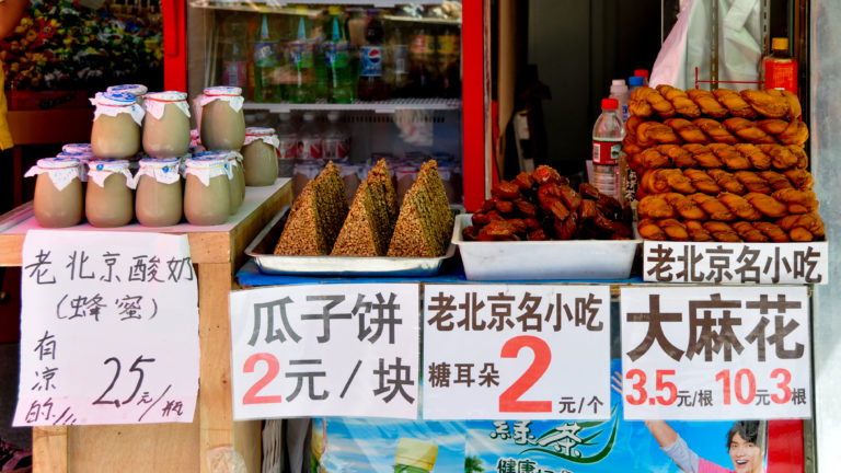 Beijing street-food stall with bottled yogurt, sesame-coated 瓜子饼 melon-seed cakes, and other snacks labeled with handwritten yuan prices.