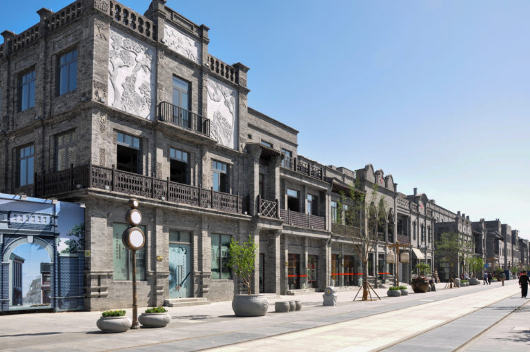 Restored gray-brick buildings with repeating balconies and decorative panels along a wide pedestrian street with modern lamps and planters.