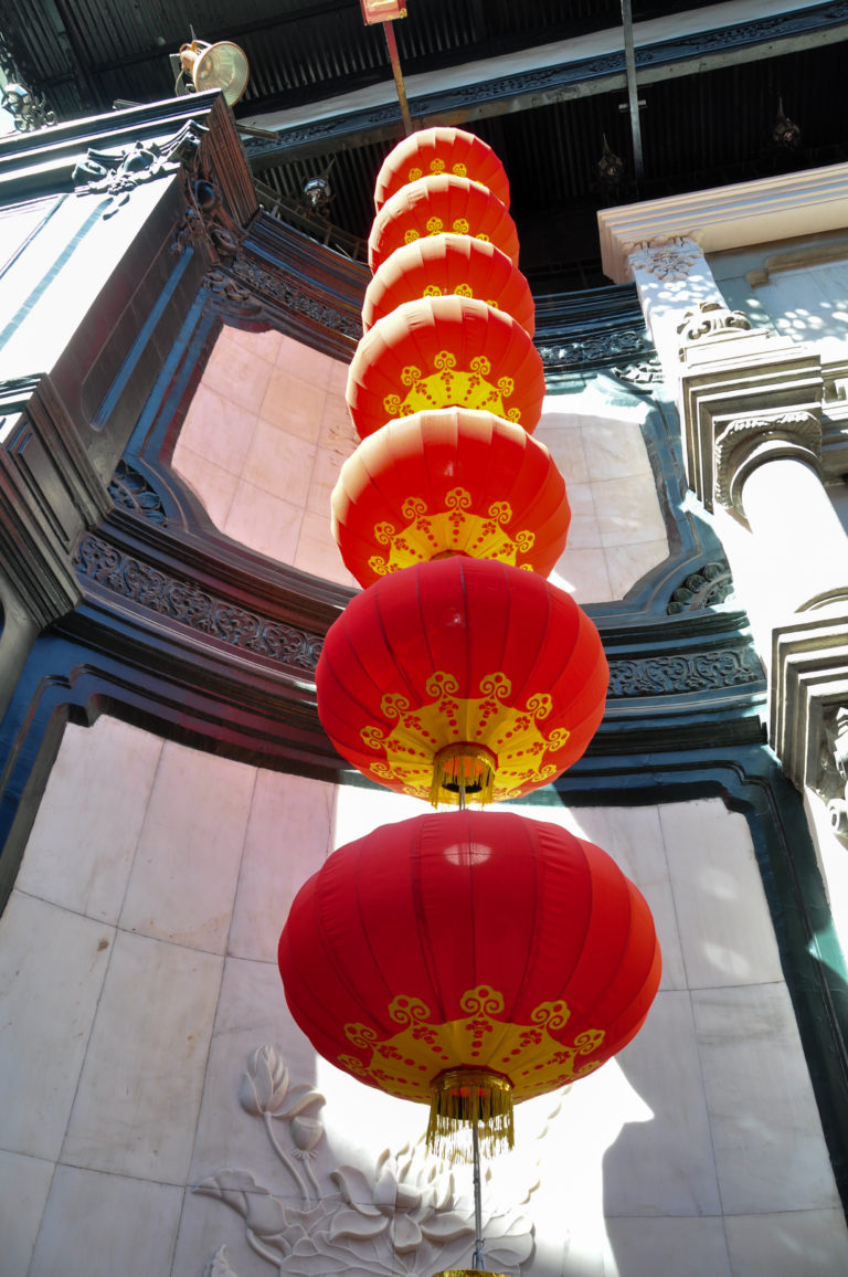 Upward view of large red lanterns with gold patterns hanging beneath curved gray cornices in a traditional Beijing courtyard.