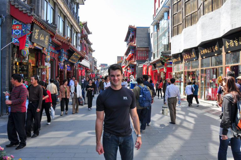 Traveler standing and facing the camera on a crowded Beijing pedestrian shopping street lined with traditional-style storefronts and red lanterns.