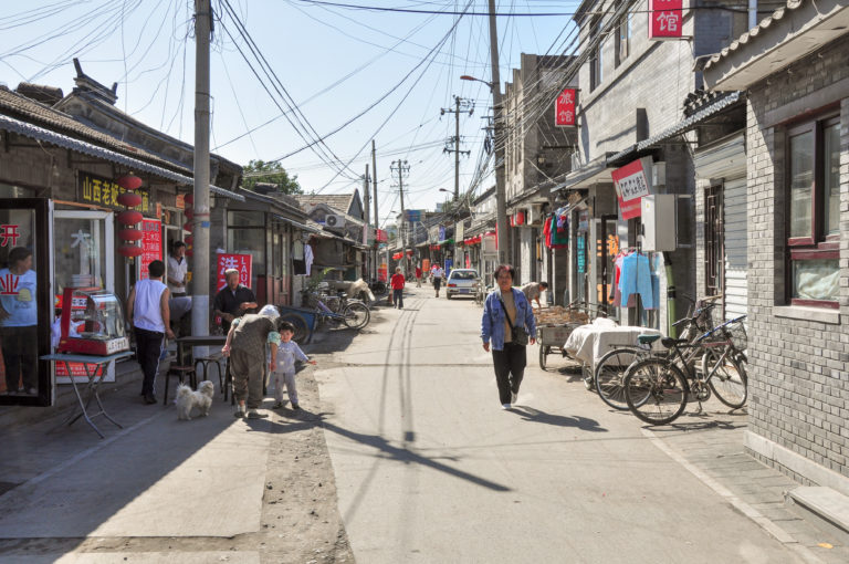 Narrow gray-brick hutong lane in Beijing with low buildings, small shops, bicycles, pedestrians, and overhead utility wires.