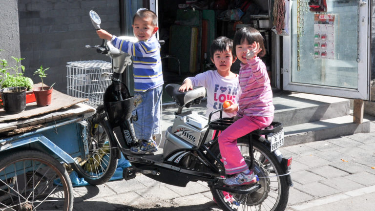 Three neighborhood children pose on parked electric bicycles outside a shop in a Beijing shopping lane.