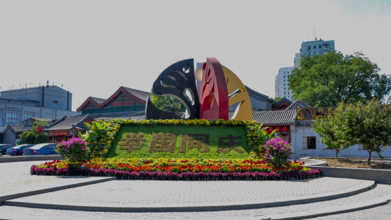Multicolored abstract metal sculpture behind a flower bed with Chinese characters formed in greenery in a Beijing public space.