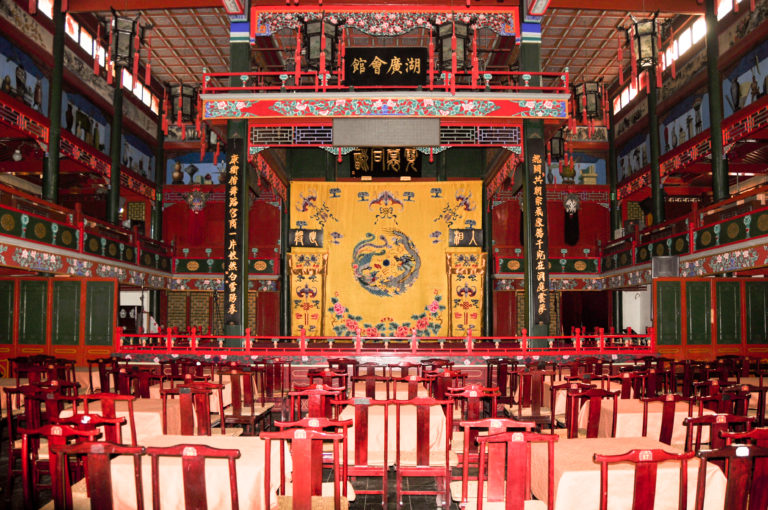 Empty traditional Chinese performance hall in Beijing with rows of red chairs facing an ornate opera stage and large yellow dragon-themed hanging.