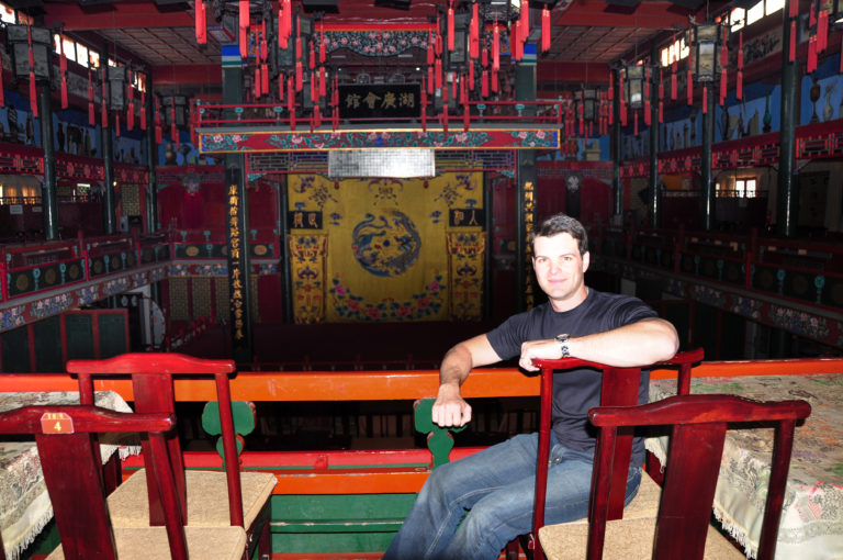 Traveler seated among red chairs facing an ornate Chinese opera stage with painted beams and a yellow embroidered backdrop in Beijing.