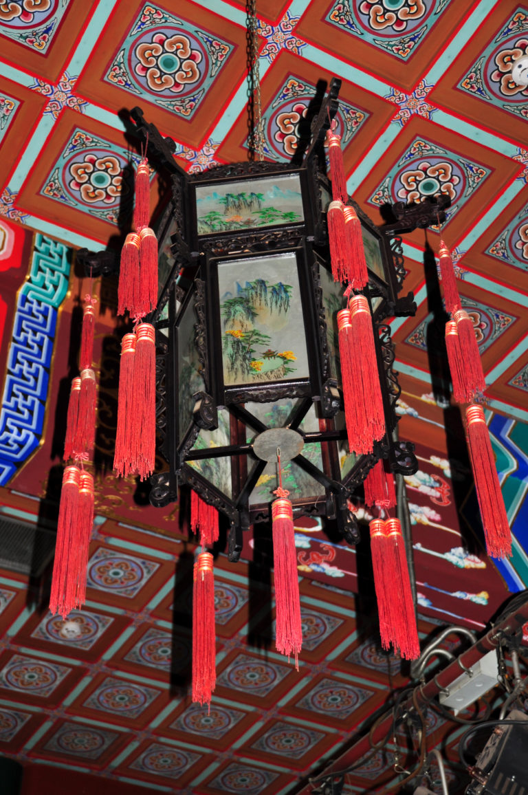 Black-framed hanging lantern with painted mountain-and-water panels surrounded by long red tassels.