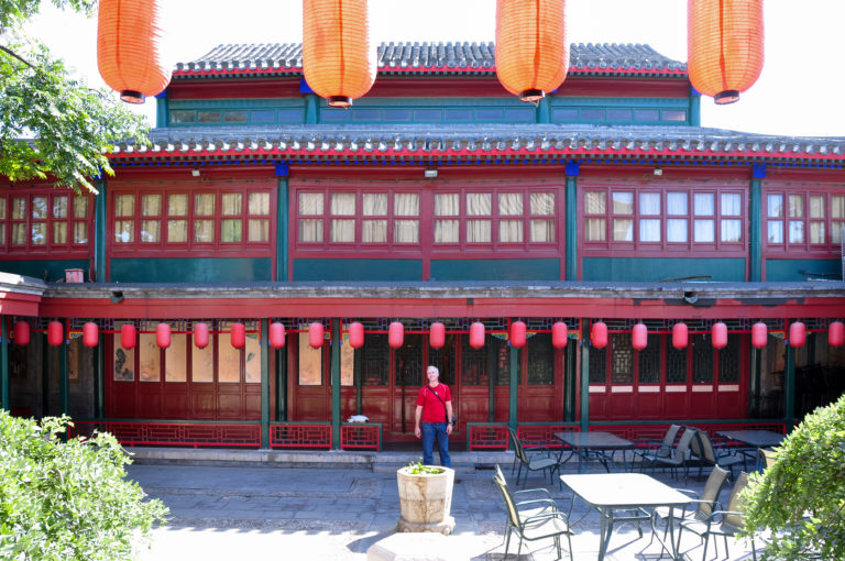 Two-story red-and-green traditional Chinese timber building with large hanging lanterns overlooking a courtyard with seating in Beijing.