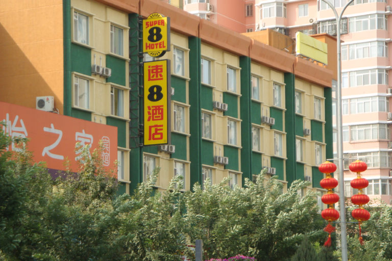 Facade of a green-and-tan Super 8 Hotel (速8酒店) in Beijing with vertical bilingual signage, rows of windows and air conditioners, and red lanterns.