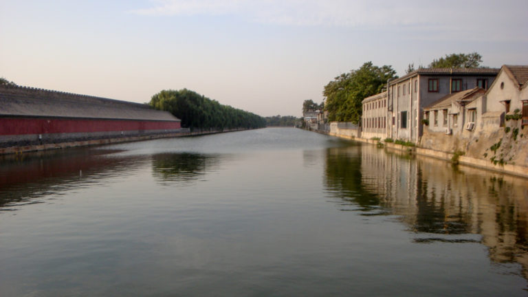 Long view of the Forbidden City moat between a red crenellated palace wall and gray low-rise buildings in Beijing.