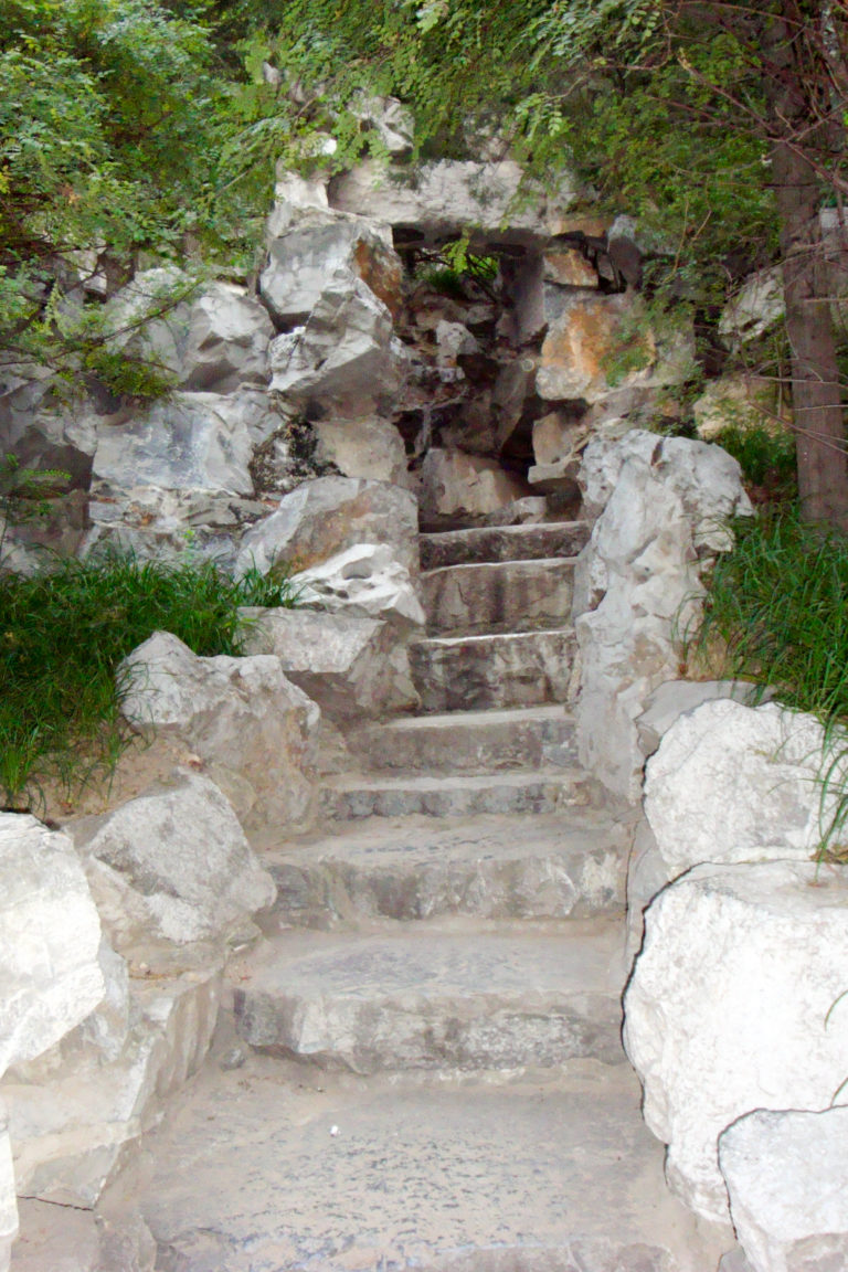 Narrow, rough stone steps rising between pale jagged rocks and vegetation in a Chinese garden rockery.