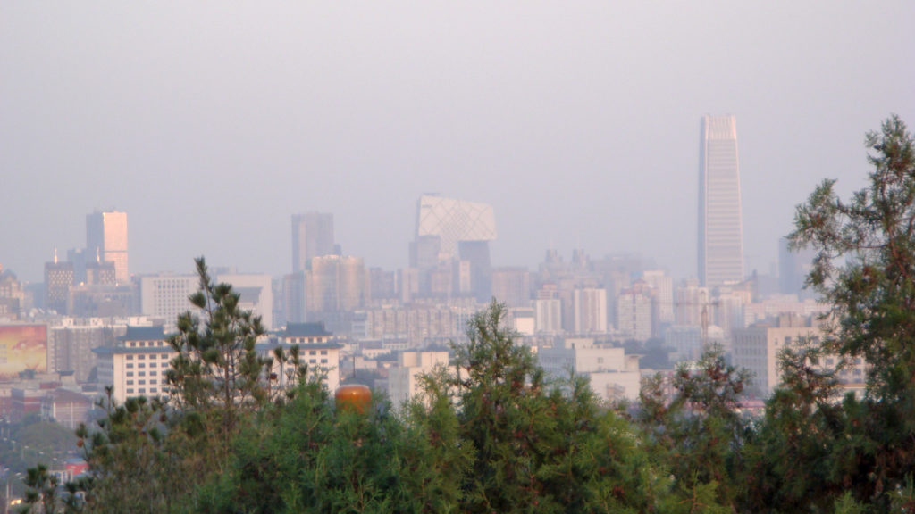 Hazy view of Beijing’s central business district skyline with the distinctive looped CCTV Headquarters and other modern high-rises beyond trees.