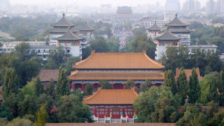 View north from Jingshan over golden-roofed traditional halls and pavilions aligned toward the Drum Tower corridor, with modern buildings fading into the distant haze.