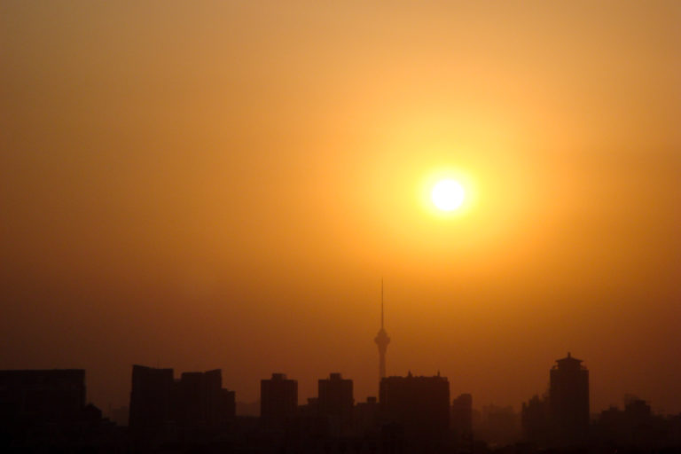 Orange sun setting in hazy sky above Beijing skyline with a communications tower and mix of high- and low-rise buildings.