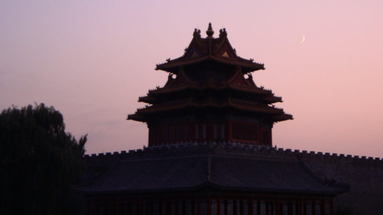 Forbidden City corner tower at dusk, silhouetted above the palace wall with layered upturned roofs and a crescent moon in a pink sky.