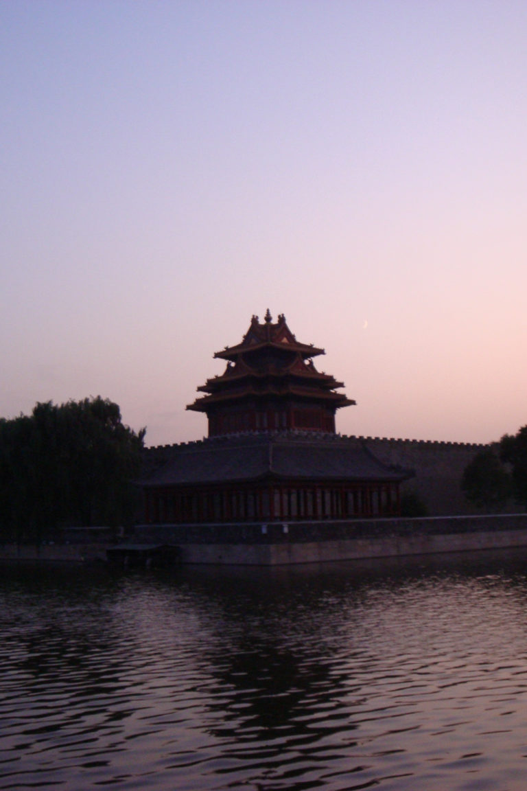 Northwest or northeast corner tower of the Forbidden City at dusk, silhouetted above the palace wall with a crescent moon in a pink evening sky.