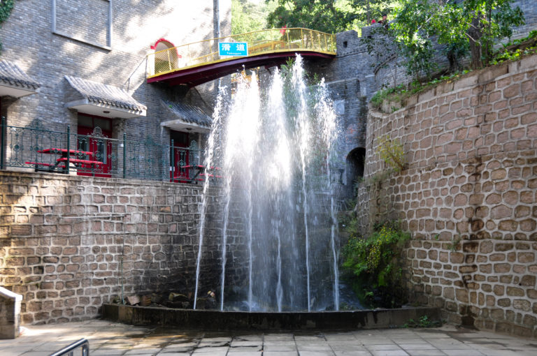 Urban waterfall dropping from a bridge into a small paved basin, framed by gray stone walls and red traditional-style architectural details.
