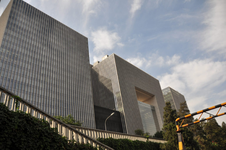 Monumental stone-and-glass Beijing office complex with deep vertical fins and a tall rectangular portal cut through the central block.