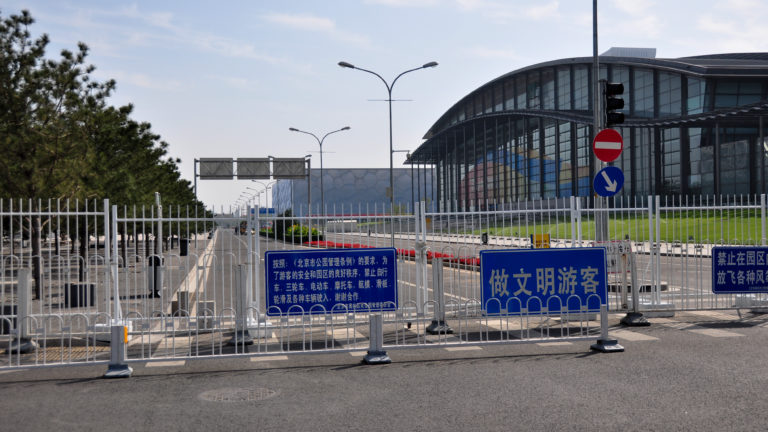 Temporary metal barriers with blue visitor-conduct signs block a road leading toward a curved glass-and-steel Olympic venue in Beijing.