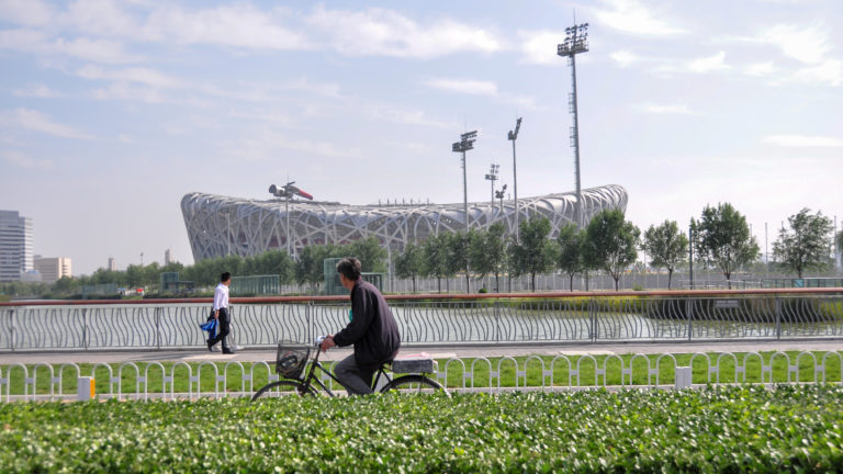 Cyclist riding past a landscaped verge with Beijing National Stadium’s nest-like steel lattice structure dominating the background.