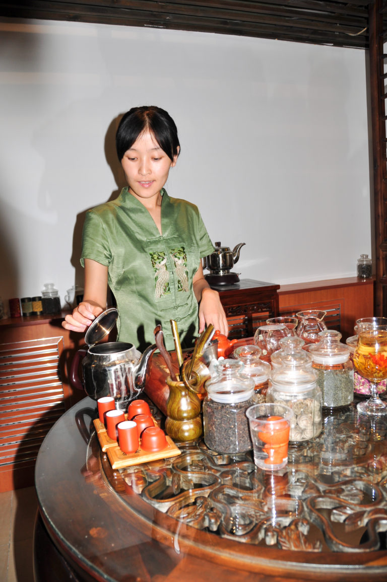 Tea attendant in a green blouse pouring from a metal kettle among glass jars of dried tea ingredients, teapots, and cups at a Chinese tea demonstration in Beijing.