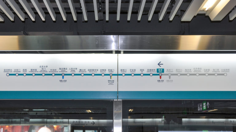 Bilingual Beijing subway route map above platform screen doors, showing station order and an arrow indicating direction of travel.