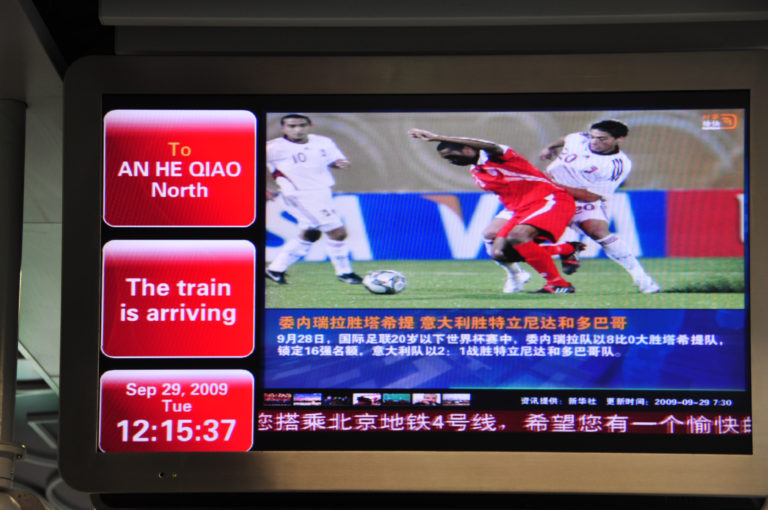 Beijing subway information screen showing a northbound train to Anheqiao North with bilingual text, date, time, and a sports broadcast.