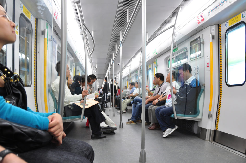Passenger view down the central aisle of a bright, modern Beijing metro train with turquoise bench seats and overhead handrails.