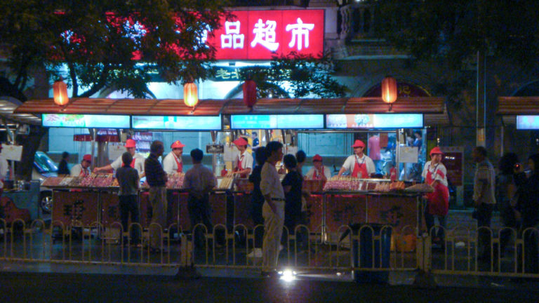 Wide nighttime view of Beijing street-food stalls with red-uniformed vendors, bright Chinese signage, red lanterns, and customers gathered at the counter.
