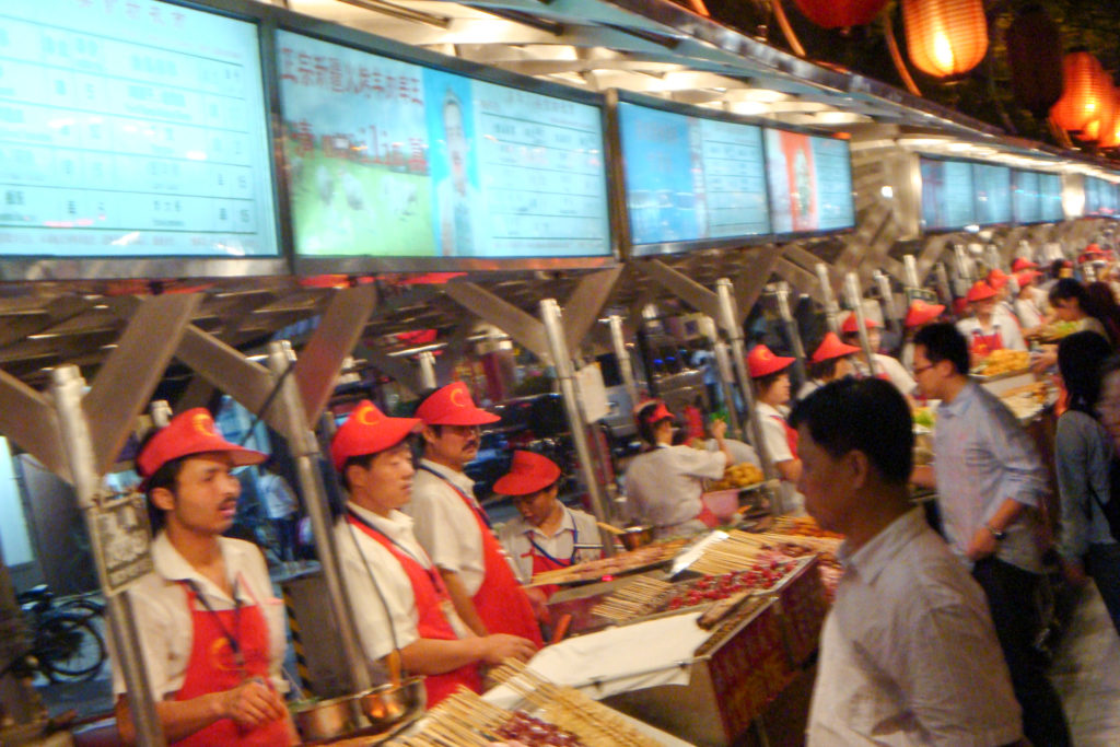 Uniformed vendors in red caps and aprons serve customers at illuminated skewer stalls in a Beijing night market.