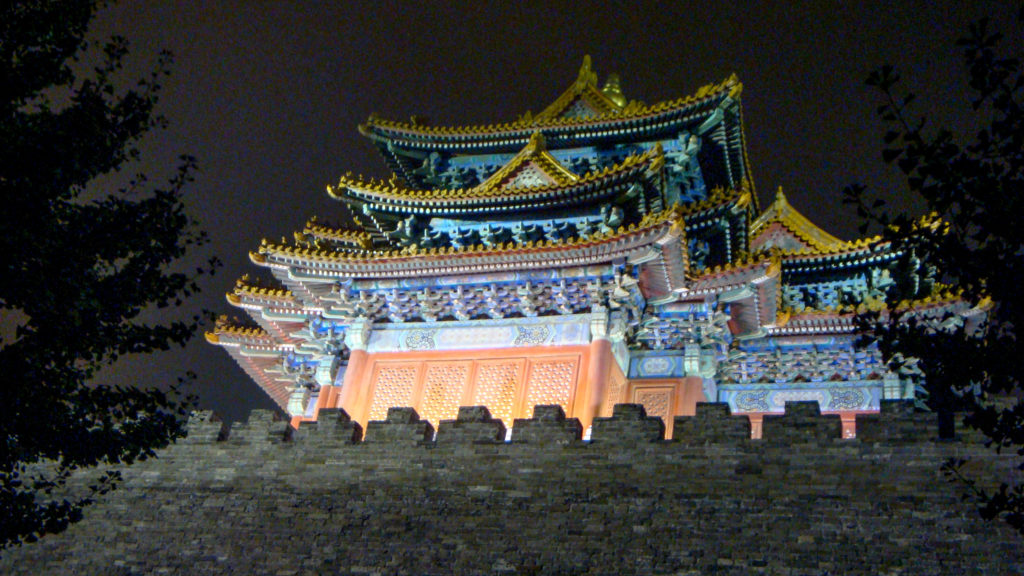 Illuminated multi-tiered traditional Chinese gate tower above a dark crenellated wall at night.