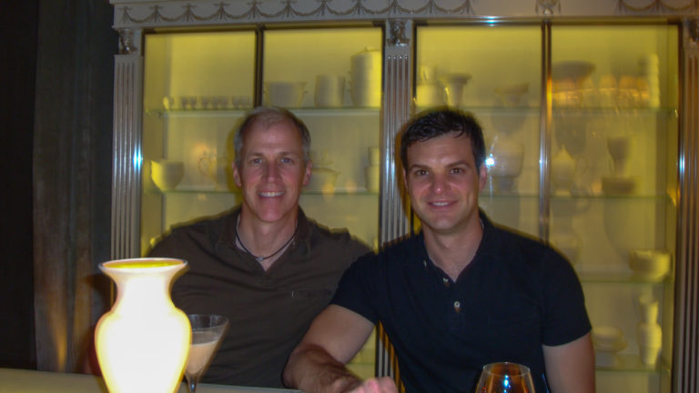 Two male travelers smiling at a table in a Beijing restaurant or bar, with a glowing lamp in the foreground and ceramic vessels on illuminated shelves behind them.