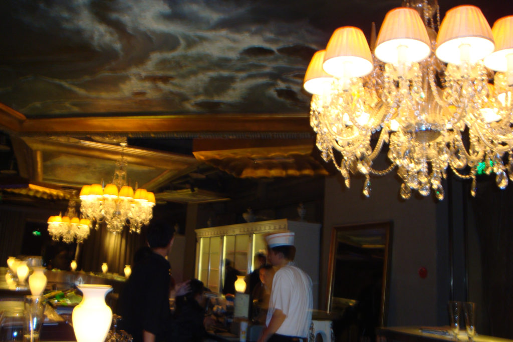 Wide interior view of a Beijing bar with multiple crystal chandeliers and warm table lamps illuminating a long counter.