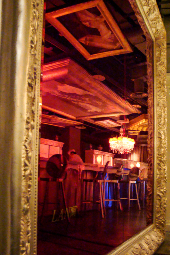 Ornate gilded mirror reflecting a red-lit bar interior with a chandelier and seated patrons at night.