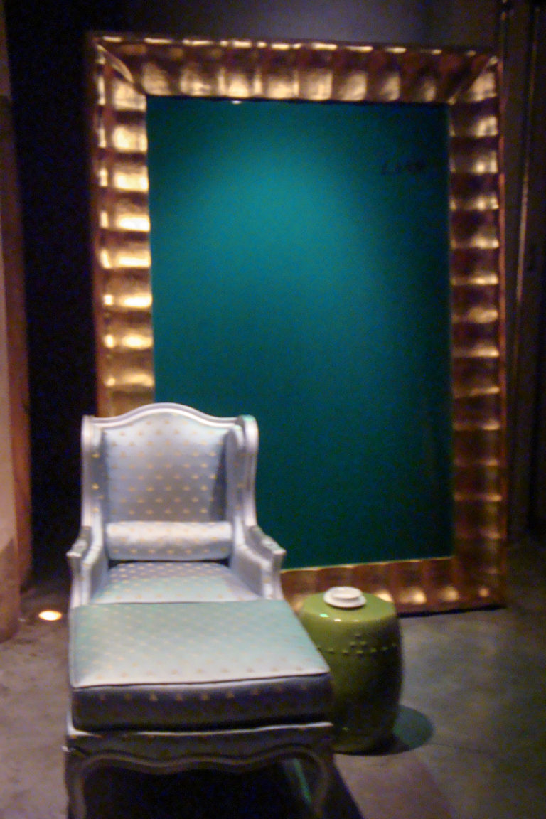 Ornate pale upholstered armchair centered against a deep green wall with vertical illuminated elements on either side.