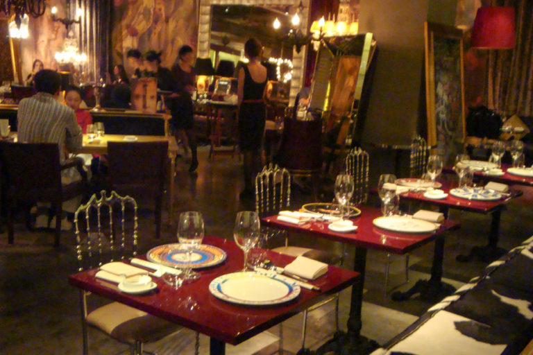 Wide view of a Beijing restaurant interior with glossy red tables, white plates, wine glasses, ornate metallic chairs, dark flooring, and diners and servers in the background.