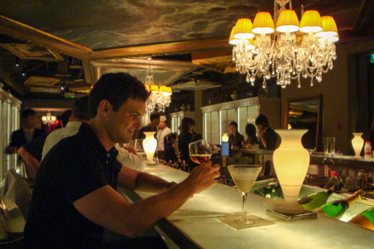 Traveler in a dark shirt sits at an illuminated bar counter in an upscale Beijing cocktail bar, with a martini glass on the counter and crystal chandeliers overhead.
