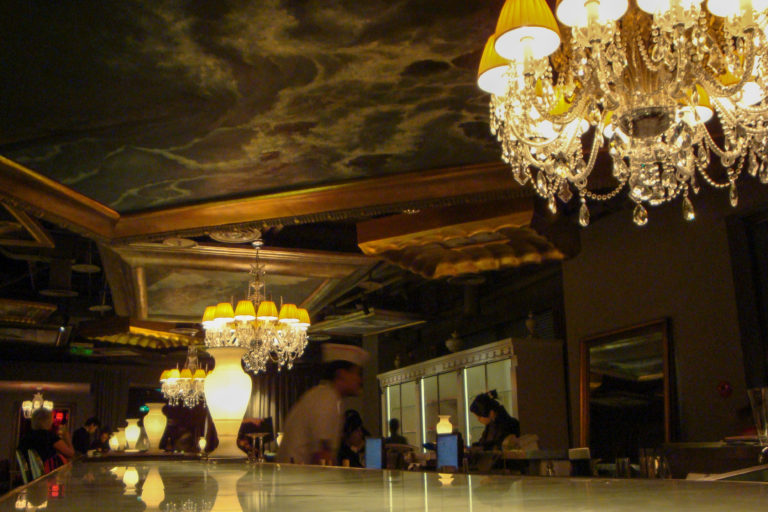 Wide view of a dark, luxurious Beijing bar with a glossy illuminated counter and multiple crystal chandeliers receding into the distance.