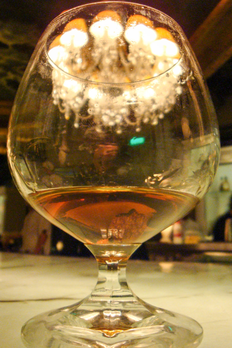 Extreme close-up of a rounded stemmed glass holding a shallow amber liquid, its curved bowl reflecting distorted lights and people in a bar.