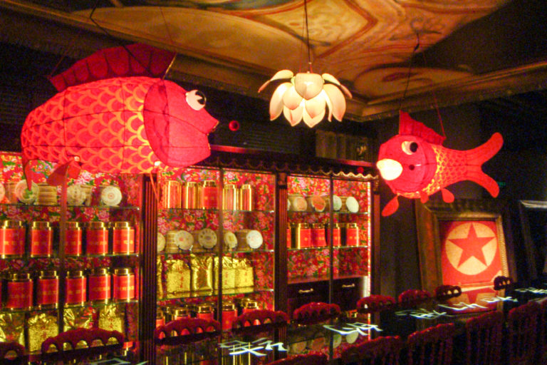 Wide view of a Beijing restaurant interior with large red fish-shaped lanterns hanging from the ceiling above glass cabinets and red seating.