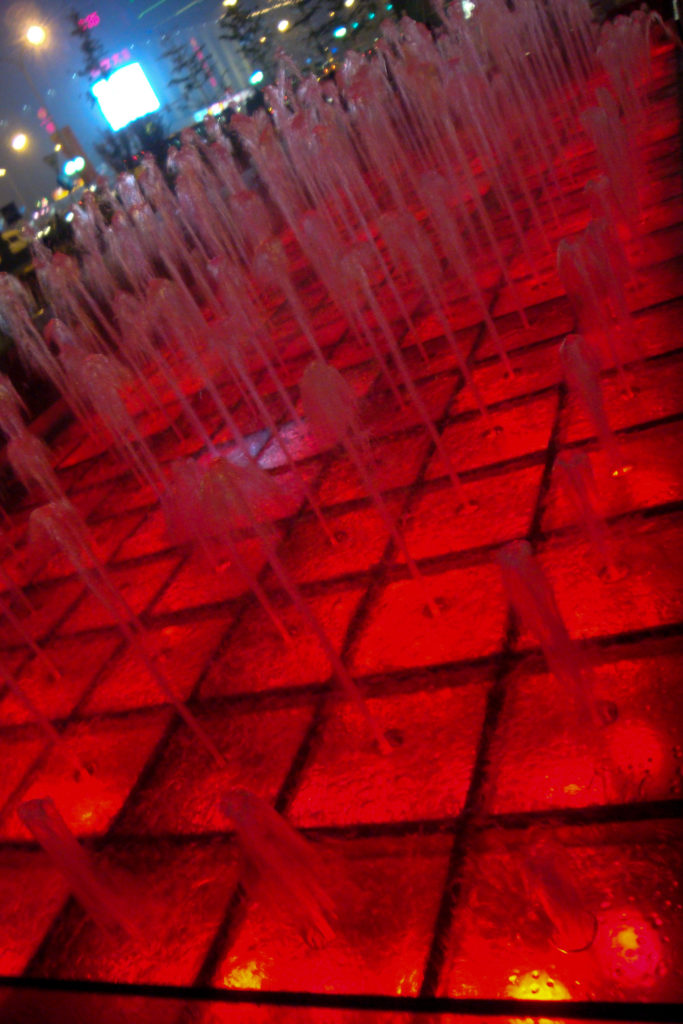 Tilted close-up of red-lit fountain grid with softened water jets forming vertical lines over square divisions.