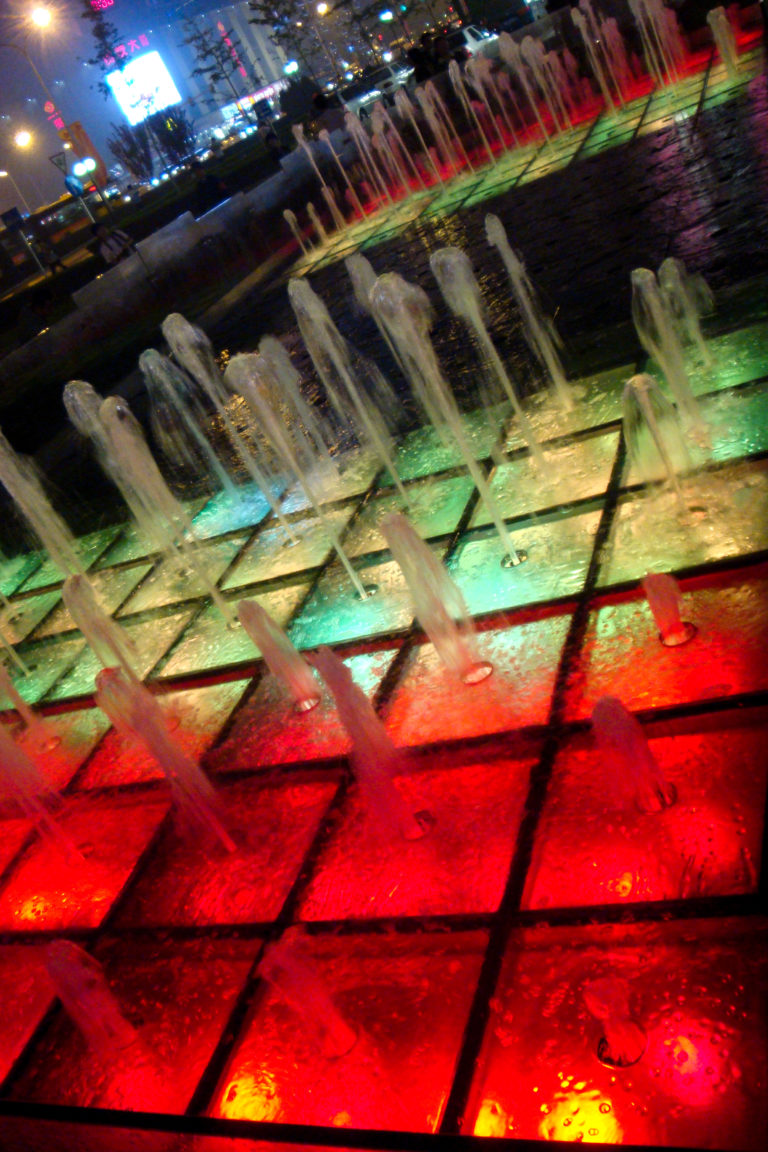Close-up of a night fountain in Beijing with vertical jets rising from a square grid, lit by red and green underwater lights.