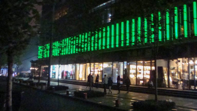 Wide night street view of a long commercial building in Beijing with vertical green LED lights across the upper facade, bright shops below, and pedestrians walking along the sidewalk under silhouetted trees.